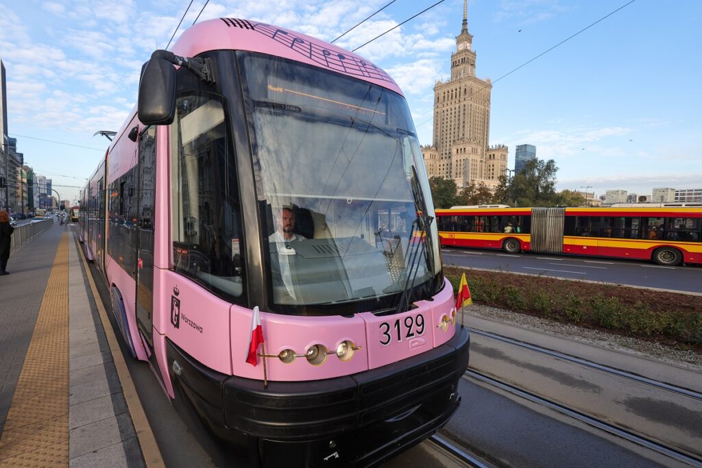 Różowy tramwaj zatrzymany na przystanku, w tle widać charakterystyczny wieżowiec oraz czerwono-żółty autobus.