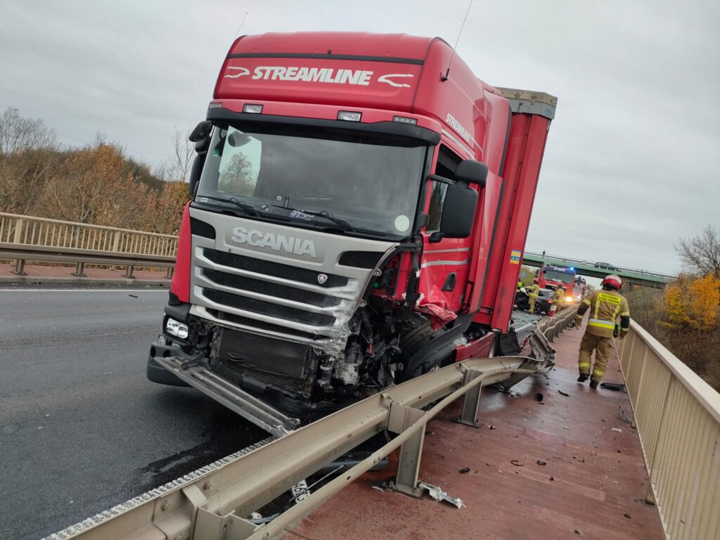 Czerwony samochód ciężarowy marki Scania z uszkodzoną kabiną stoi na moście, obok pracują strażacy w odblaskowych kamizelkach.