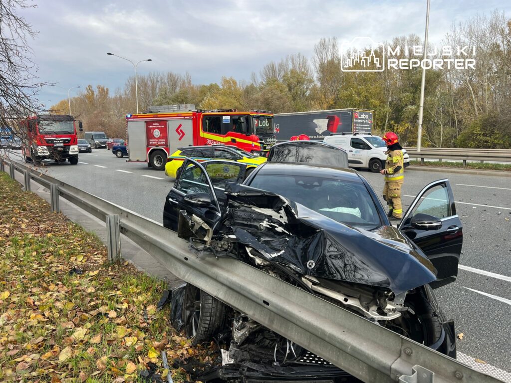 Czarny samochód osobowy z uszkodzoną tylną częścią stoi przy barierce drogi, obok interweniują strażacy i karetka.