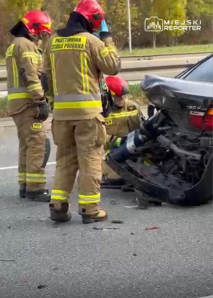 Czterech strażaków w mundurach i hełmach, pracujących przy uszkodzonym samochodzie osobowym na drodze.