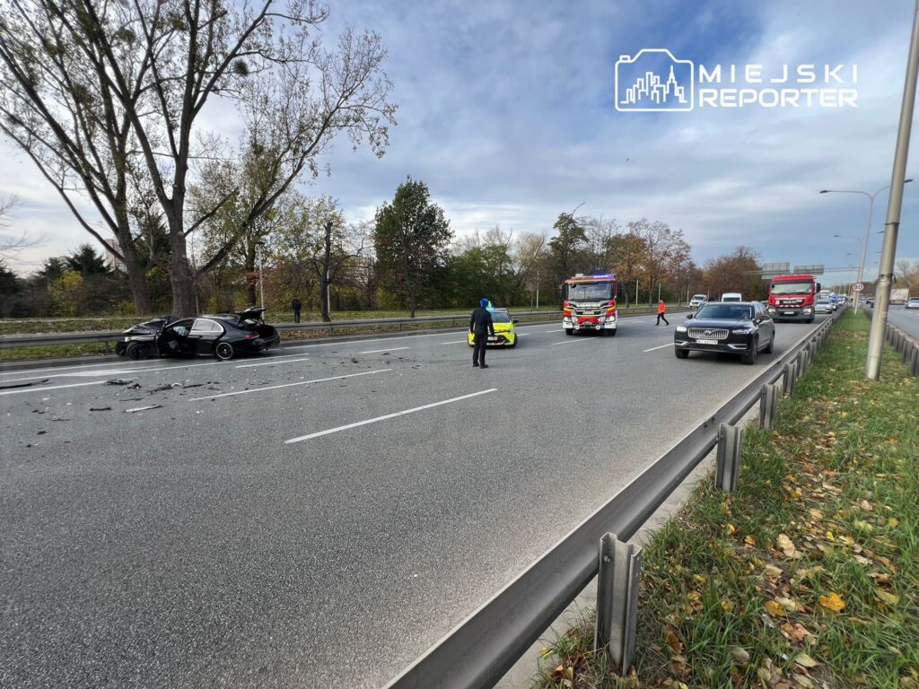 Czarny samochód osobowy z uszkodzoną tylną częścią stoi na pasie drogi, obok niego pracują strażacy i policjanci.