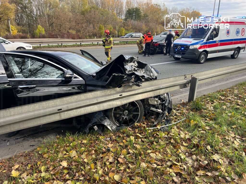 Czarny samochód osobowy z uszkodzoną przednią częścią stoi przy barierce na poboczu drogi, w tle widać karetkę i ratowników.