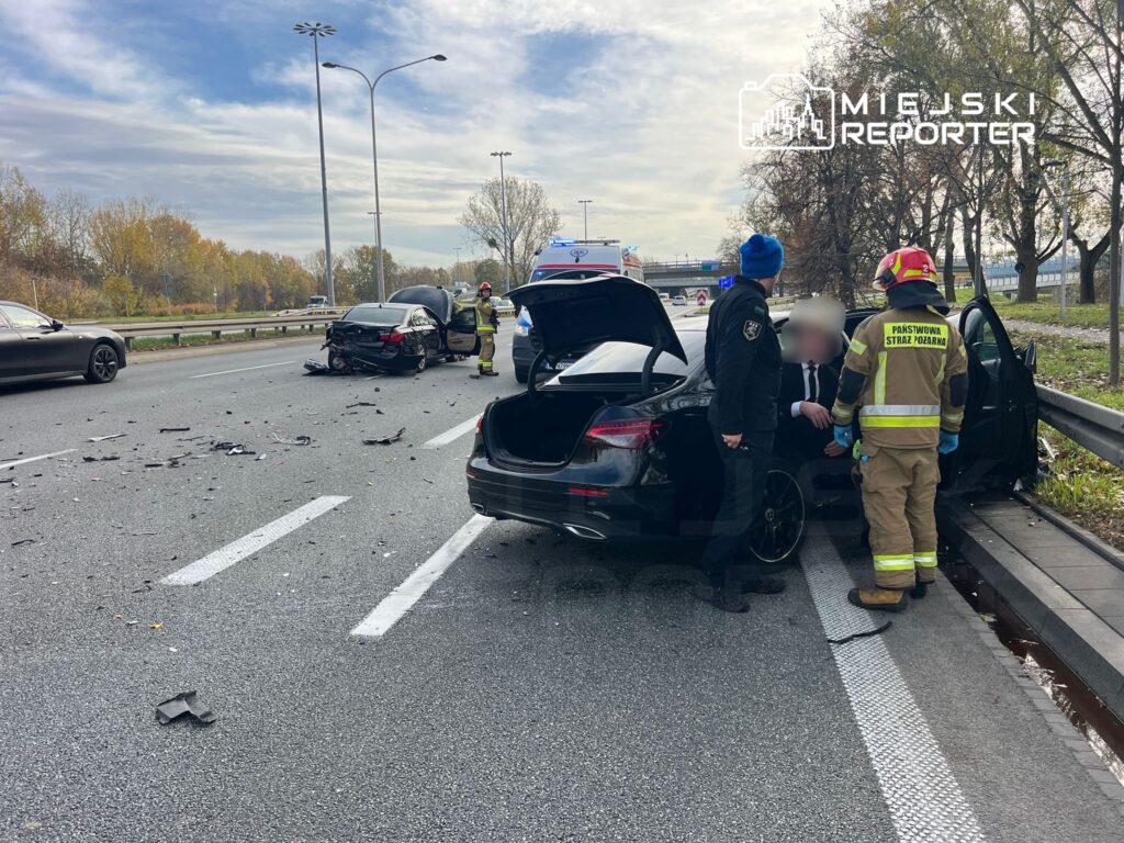 Na drodze leży rozbite auto z uszkodzoną tylną częścią, obok stoją strażacy i policjant, pomagający kierowcy.