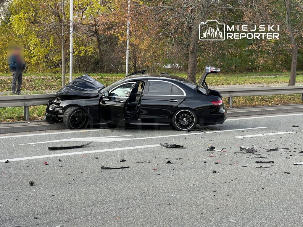 Czarny samochód osobowy z uszkodzoną przednią częścią stoi na jezdni, obok leżą odłamki i zniszczone elementy pojazdu.