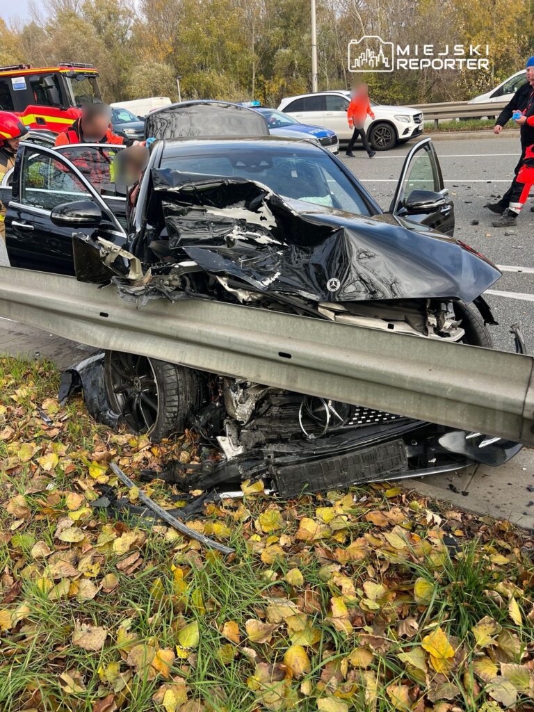 Czarny samochód osobowy z uszkodzoną przednią częścią stoi przy barierce na drodze, otoczony opadłymi liśćmi. Ratownicy udzielają pomocy poszkodowanemu.
