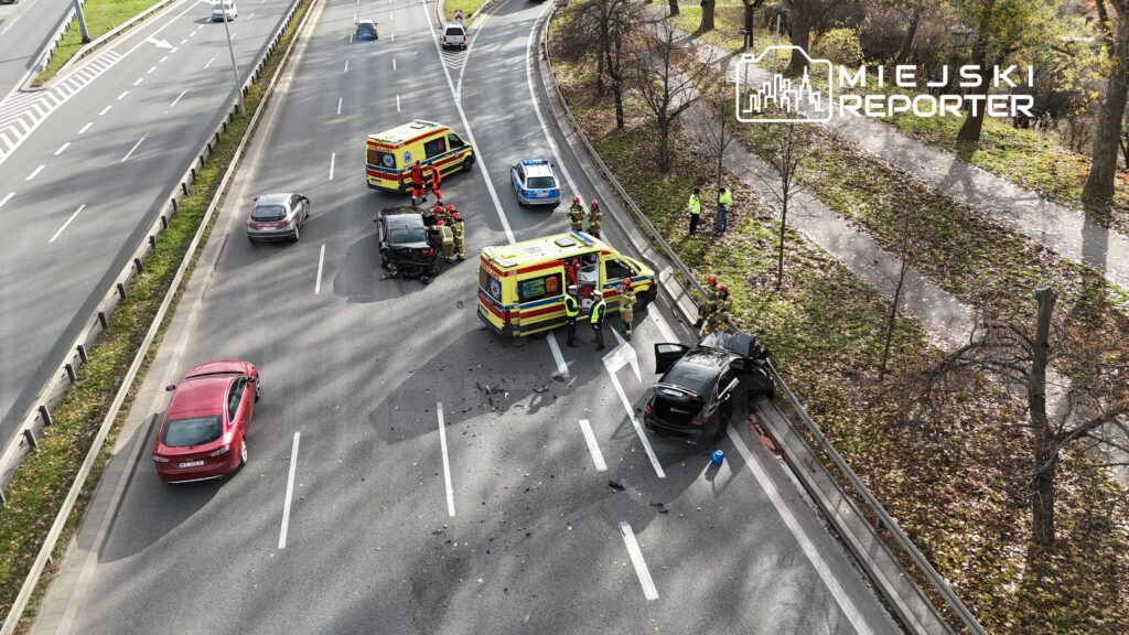 Na drodze doszło do wypadku, w którym uczestniczą dwa uszkodzone samochody; obok działają ratownicy i policja.