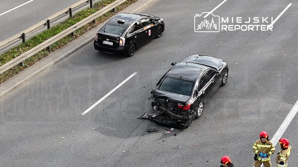 Czarny samochód osobowy z uszkodzoną tylną częścią stoi na pasie drogi, obok niego zespół strażaków w hełmach.