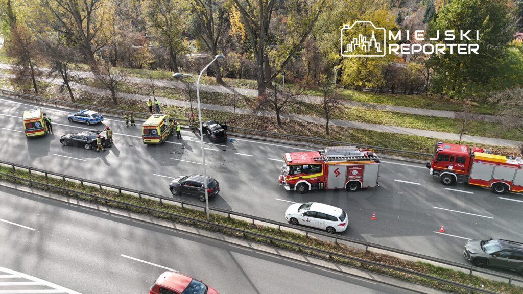 Na jezdni doszło do wypadku, przy którym pracują strażacy oraz ekipy ratunkowe; w tle widać uszkodzony samochód.