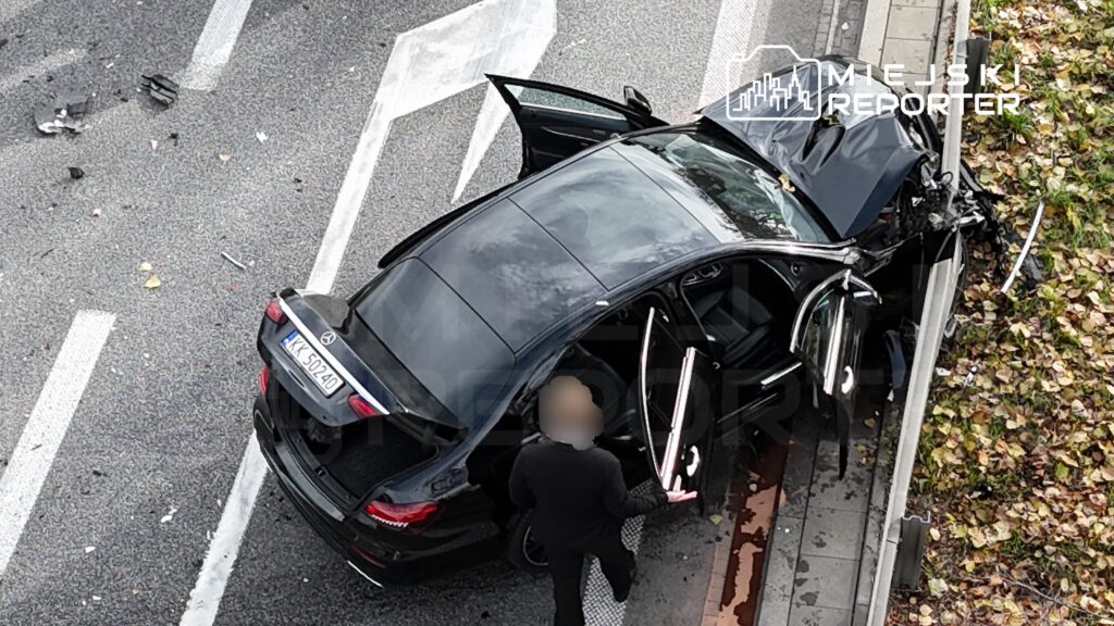 Czarny samochód osobowy z uszkodzoną karoserią stoi na jezdni, obok mężczyzna z otwartymi drzwiami wyjściowymi.