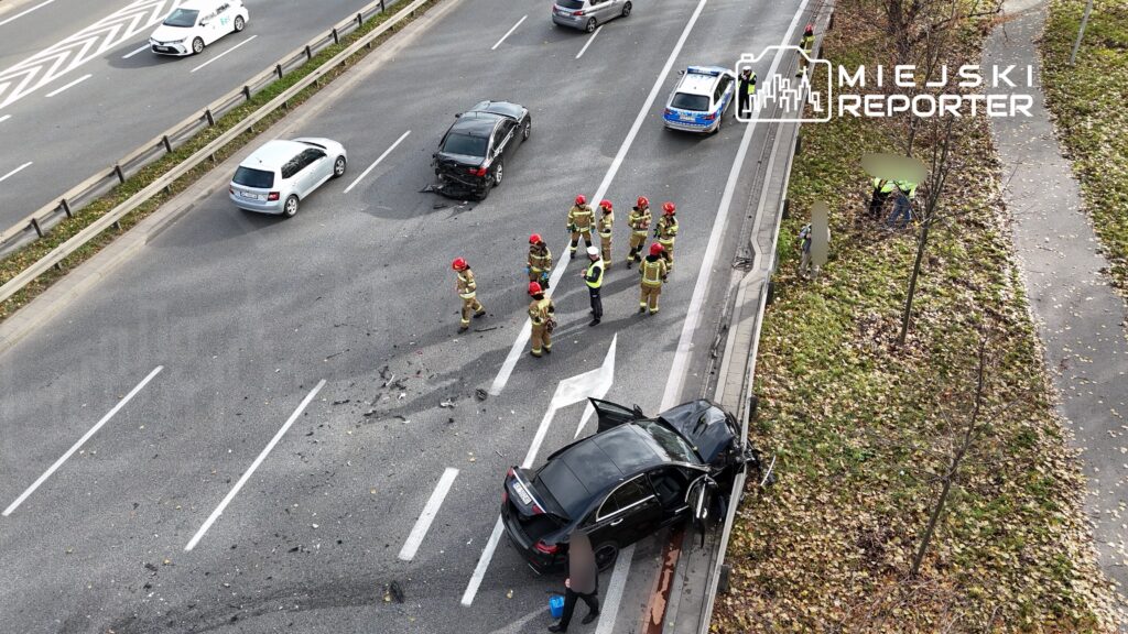 Grupa strażaków i policjantów bada miejsce wypadku samochodowego na ruchliwej drodze, przy uszkodzonym pojeździe.