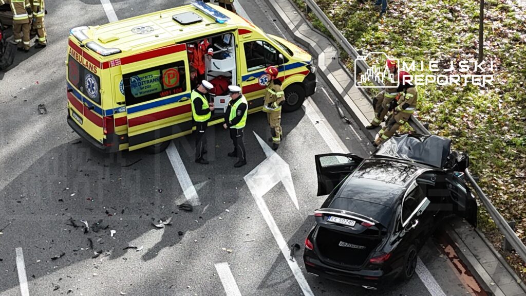 Na jezdni widać wypadek drogowy; policjanci i strażacy pracują przy czarnym samochodzie, obok stoi ambulans.