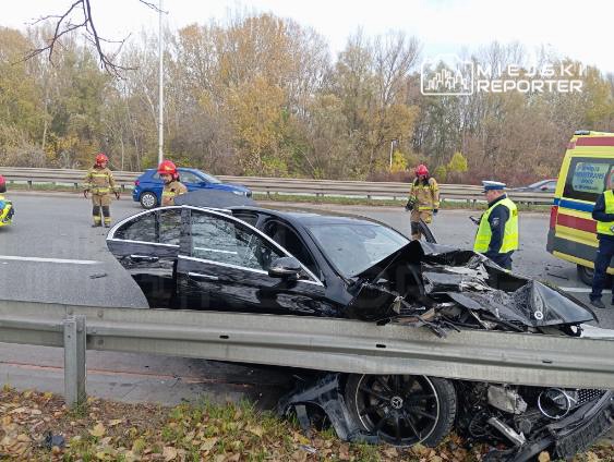 Czarny samochód osobowy zniszczony w wyniku wypadku, otoczony przez strażaków i policjantów na drodze.