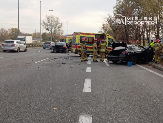 Grupa strażaków udziela pomocy poszkodowanym w wypadku drogowym na ruchliwej jezdni, obok stoi karetka.