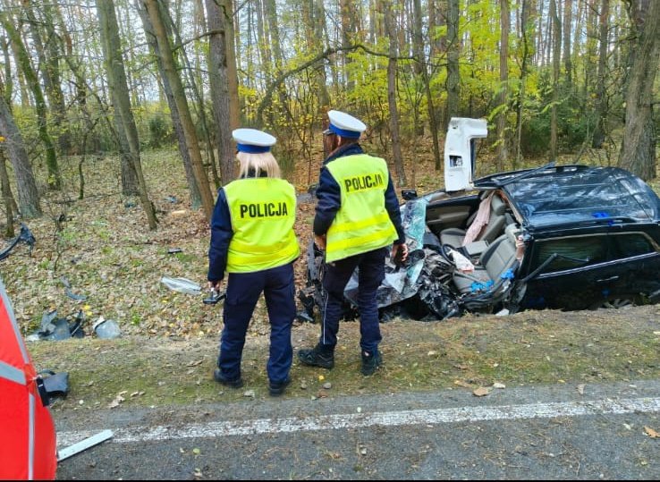 Dwóch policjantów w kamizelkach odblaskowych obserwuje wrak samochodu leżącego w rowie przy leśnej drodze.