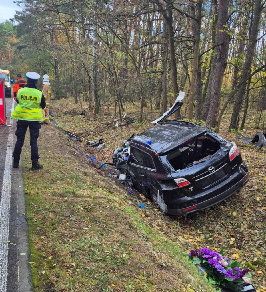 Policjant w kamizelce odblaskowej bada miejsce wypadku, obok stoi uszkodzony czarny samochód w rowie, otoczony drzewami.