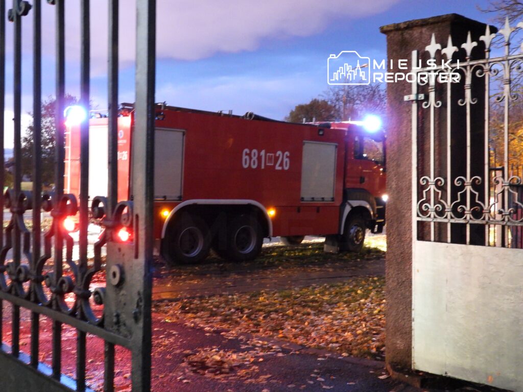 Czerwony wóz strażacki z numerem 68126 zaparkowany przy bramie, otoczony opadłymi liśćmi w wieczornym świetle.