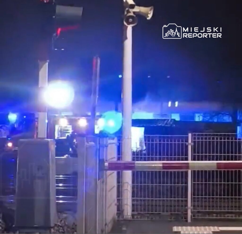 Pojazdy służb ratunkowych z niebieskimi światłami na stacji kolejowej w nocy, w tle widoczna brama kolejowa.