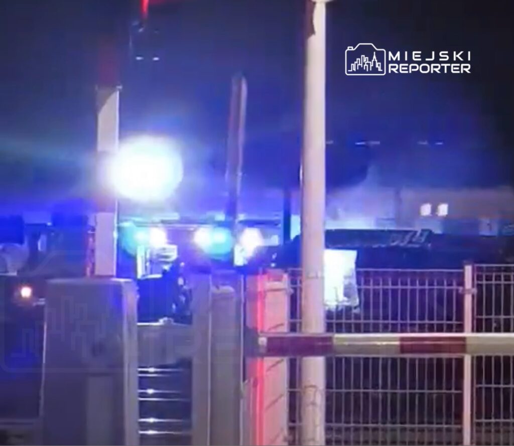 Niebieskie światła sygnalizacyjne pojazdów ratunkowych oświetlają nocną scenerię przy zamkniętej szlabanem drodze.