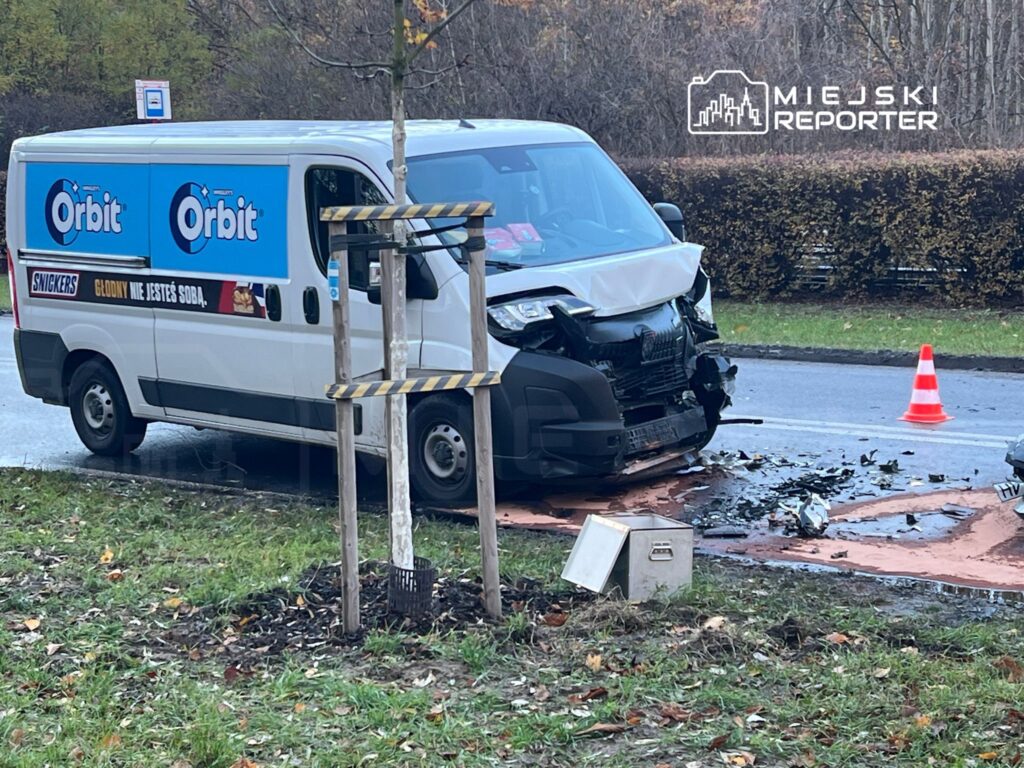 Biały samochód dostawczy z uszkodzoną przednią częścią stoi na drodze, obok leży rozlany płyn. W tle widoczne znaki drogowe i krzewy.