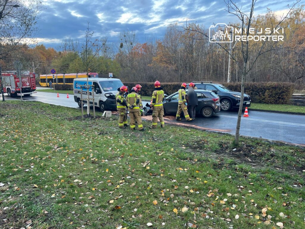 Grupa strażaków w mundurach omawia sytuację przy uszkodzonym samochodzie na poboczu drogi, obok zaparkowanej karetki.
