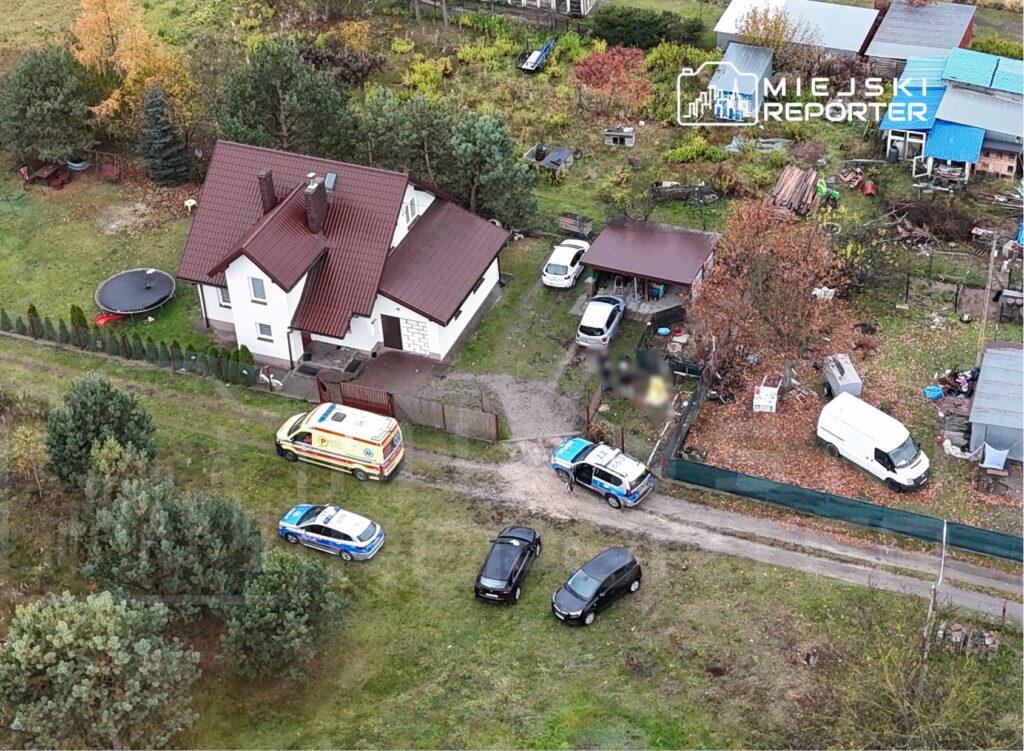 Karetka pogotowia i radiowóz policyjny na drodze przed domem jednorodzinnym w otoczeniu zieleni i samochodów.