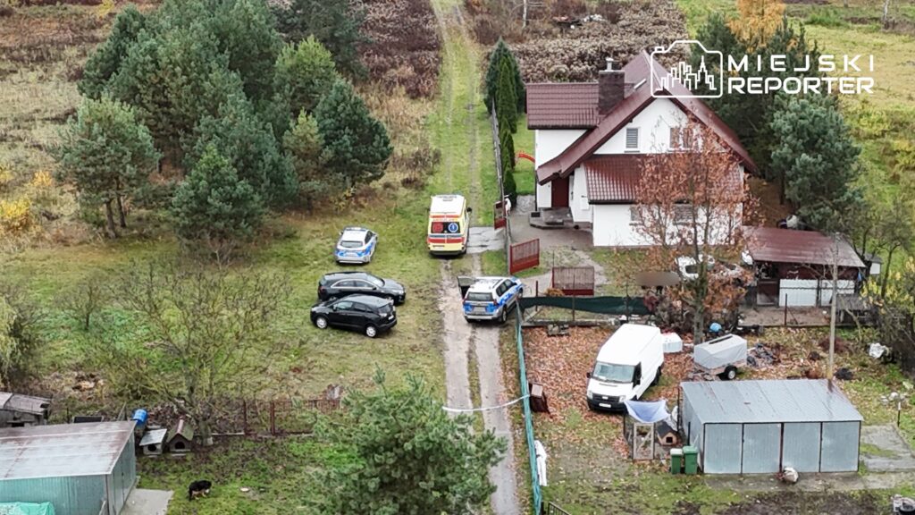 Na wąskiej drodze dojazdowej do domu stoją trzy radiowozy oraz karetka, a obok bawi się pies.