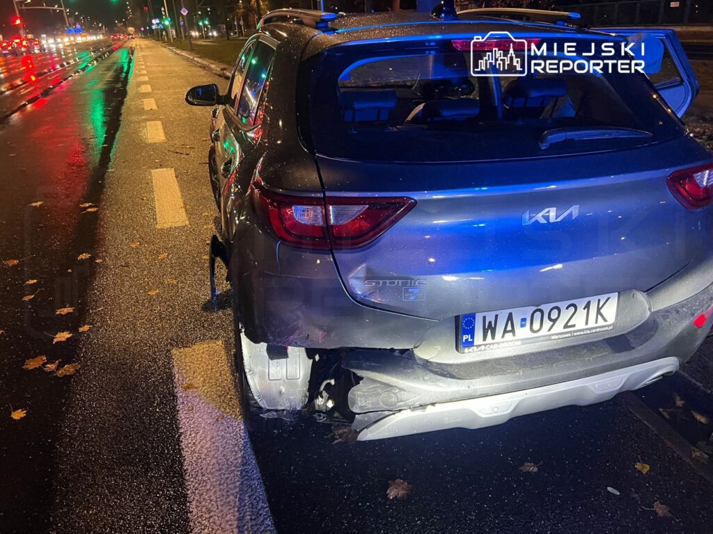 Uszkodzony samochód osobowy stojący na mokrej ulicy, w tle widać refleksy świateł i opadłe liście.