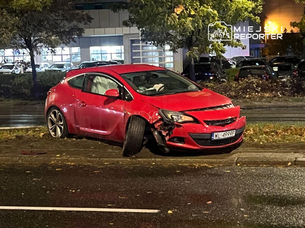 Czerwony samochód osobowy z uszkodzoną przednią częścią stoi na poboczu drogi, obok parkingu.