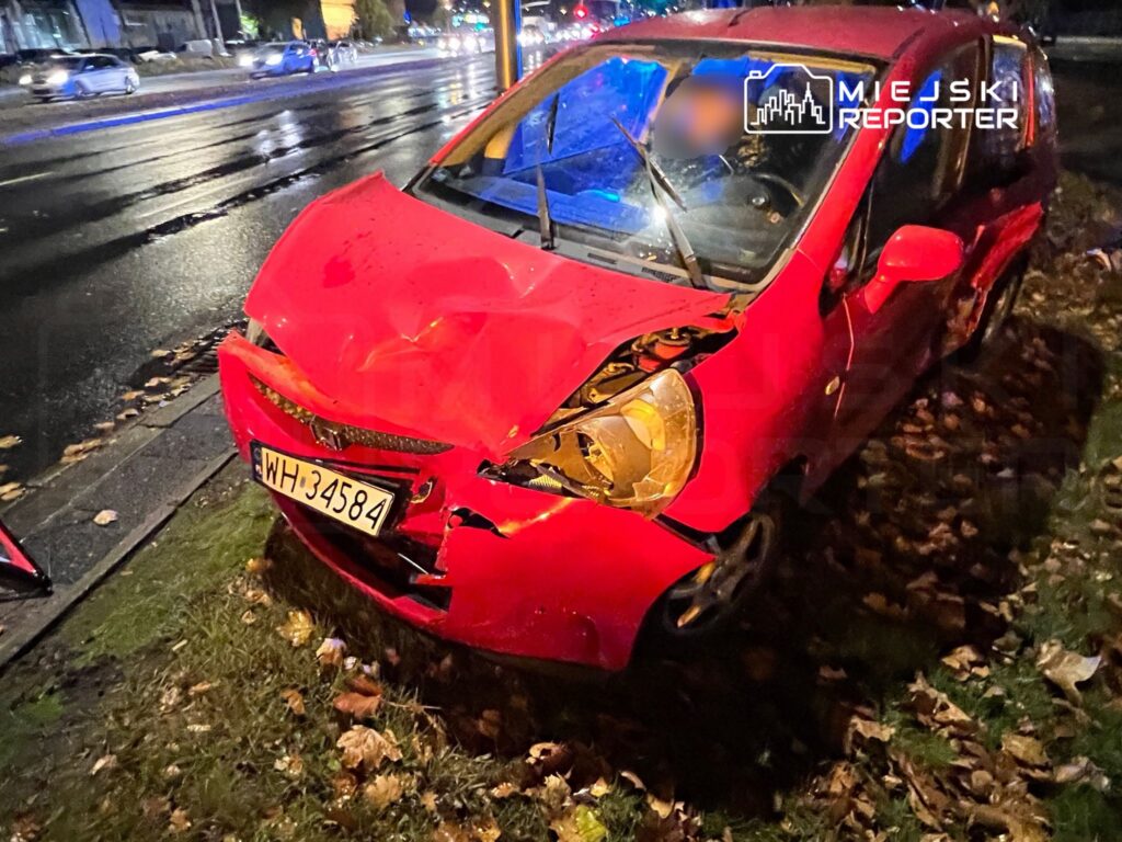Czerwony samochód osobowy z uszkodzoną przednią częścią stoi na trawie przy ulicy, obok mokrej jezdni.