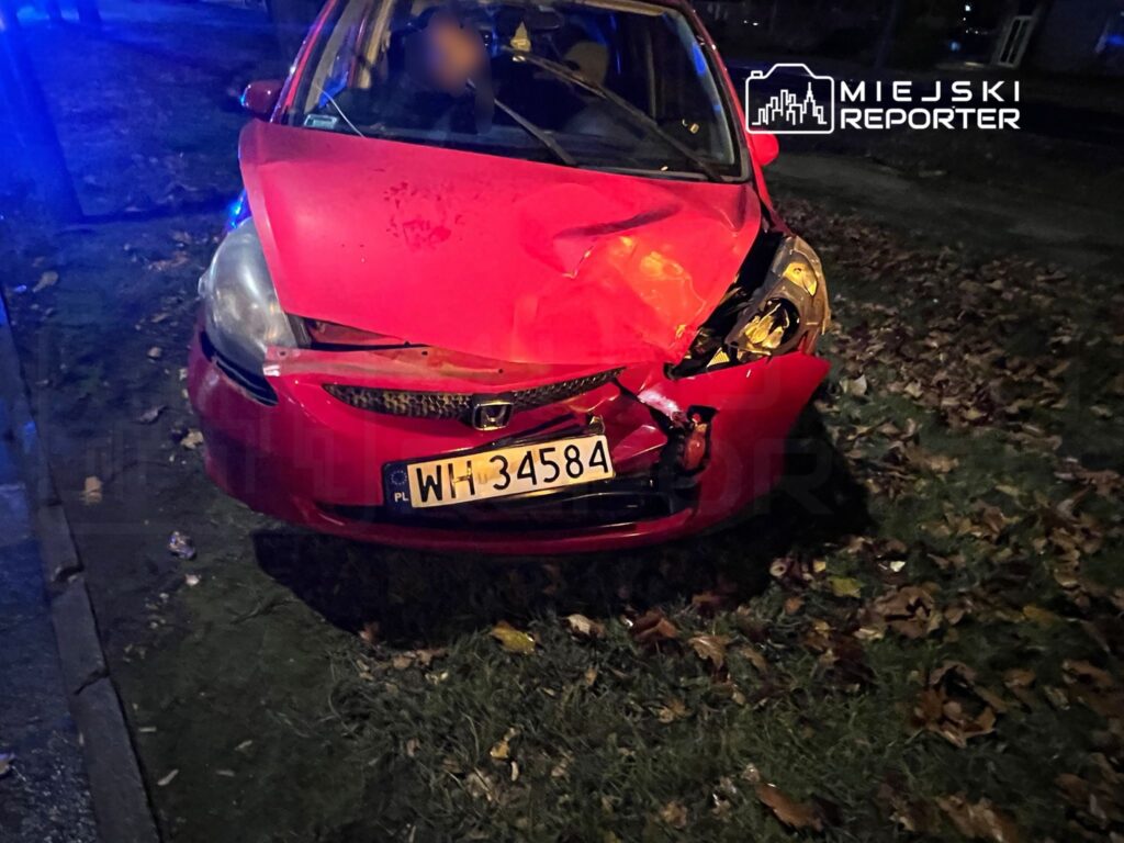 Czerwony samochód osobowy z uszkodzoną przednią częścią stoi na poboczu, w tle widać niebieskie światła.