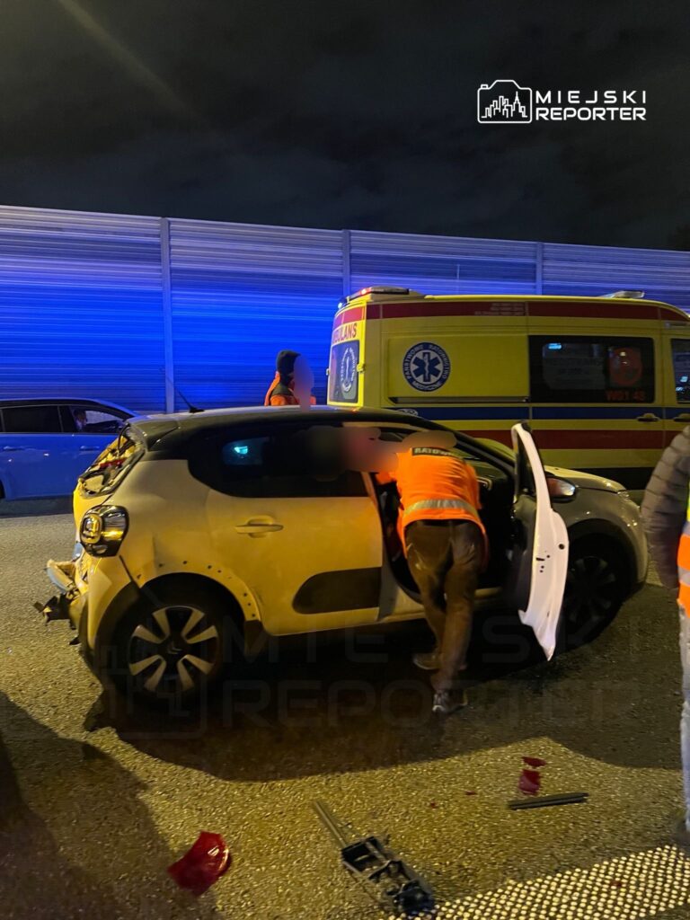 Osoby w pomarańczowych kamizelkach udzielają pomocy poszkodowanemu w wypadku drogowym przy uszkodzonym pojeździe.