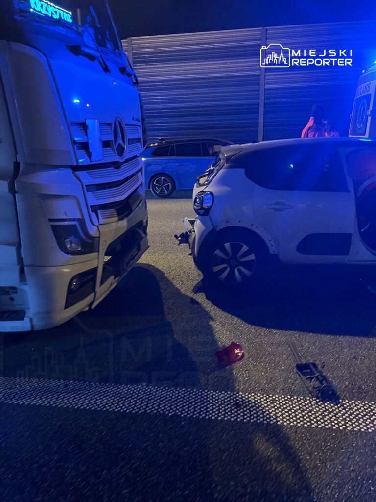 W nocy na drodze stoi ciężarówka obok uszkodzonego samochodu osobowego, a na jezdni leżą fragmenty lampy.