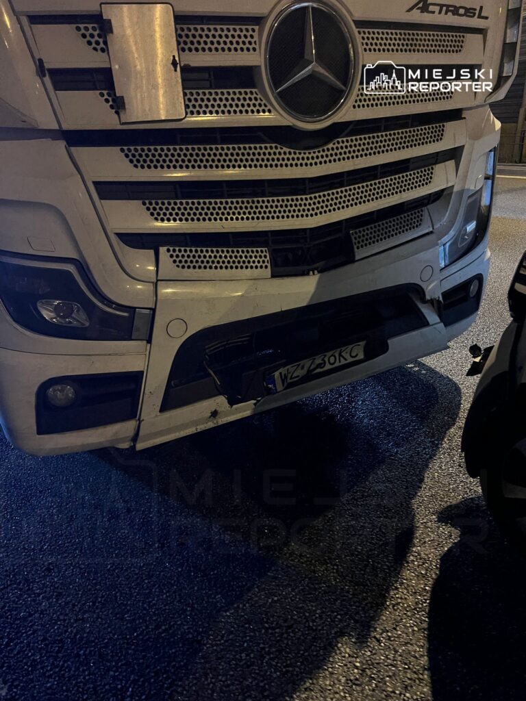 Czarny ciężarowy pojazd marki Mercedes z uszkodzonym przodem stoi na oświetlonej ulicy.