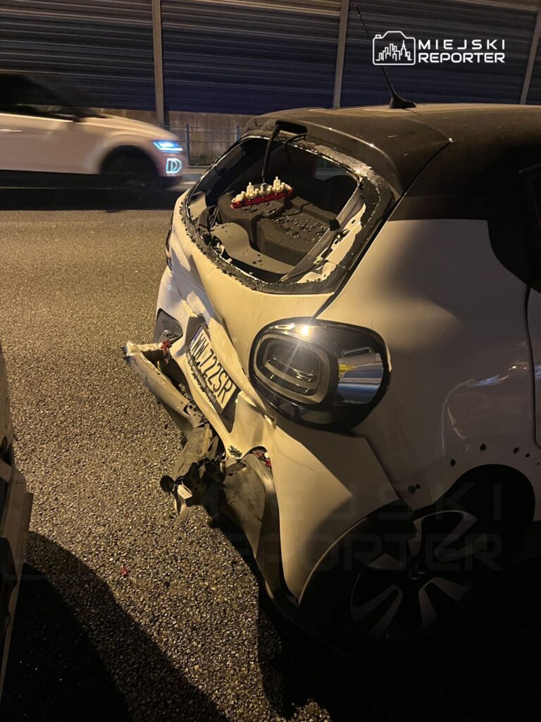 Uszkodzony biały samochód osobowy z otwartą klapą bagażnika stoi na drodze, obok przelatujący inny pojazd.