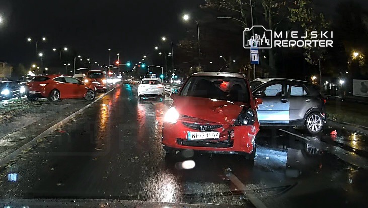 Czerwony samochód osobowy z uszkodzoną przednią częścią stoi na mokrej jezdni, obok niego szary pojazd z otwartymi drzwiami.