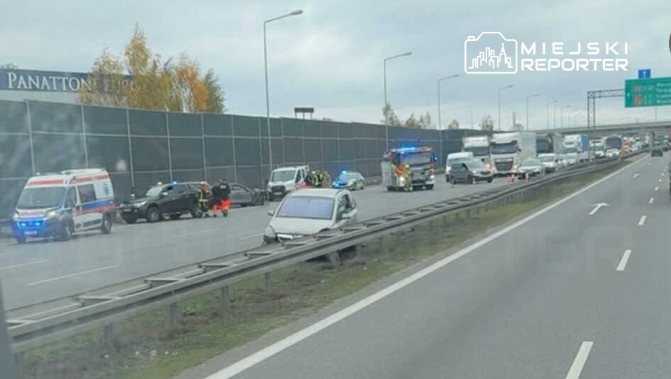 Na drodze widoczne są pojazdy służb ratunkowych oraz policyjnych, które interweniują przy wypadku. W tle zator drogowy.