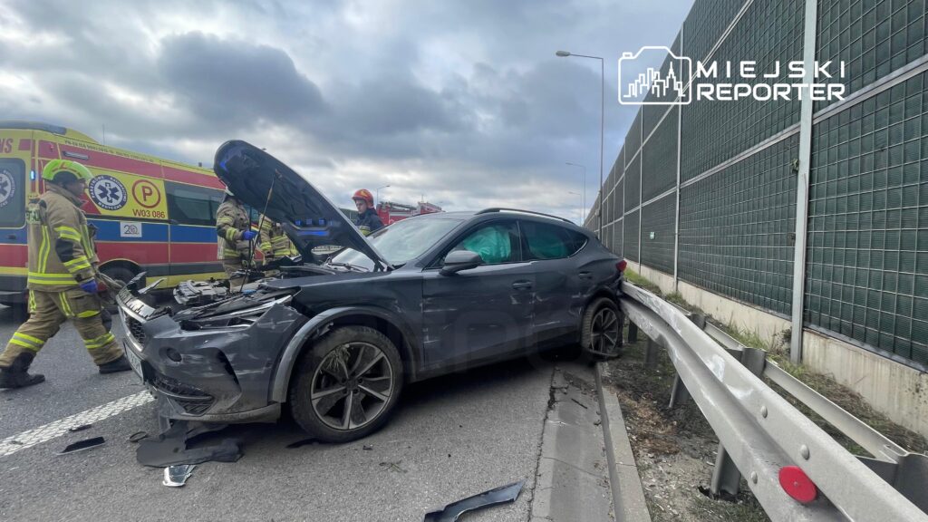 Czarny samochód osobowy z uszkodzoną przednią częścią stoi przy barierze ochronnej, obok pracują strażacy.