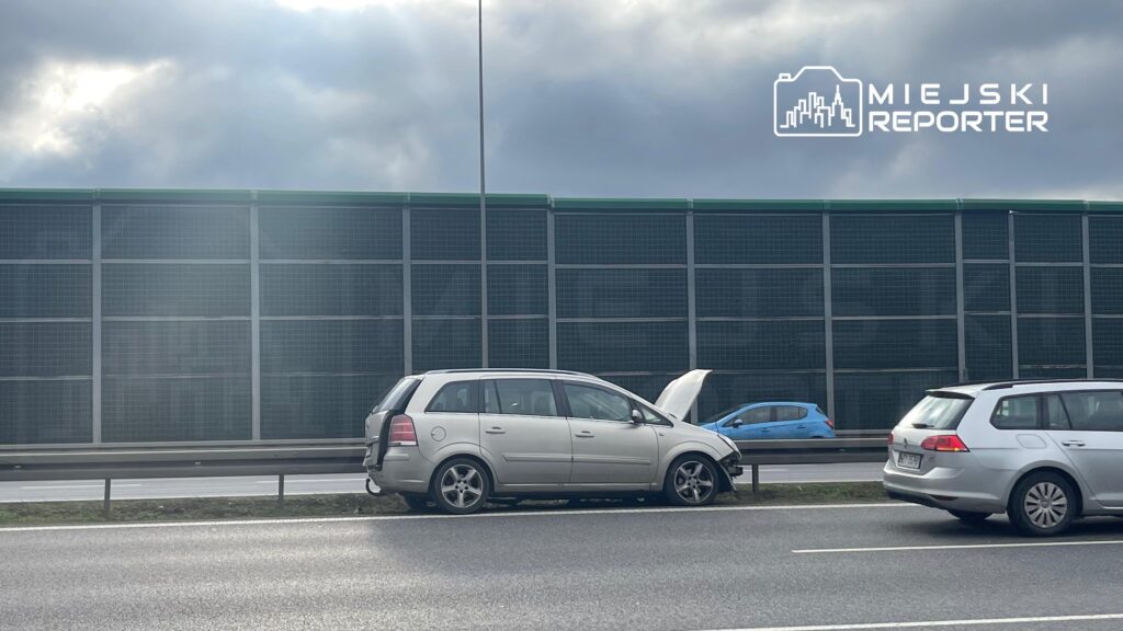 Osobowy samochód z otwartą maską stoi na poboczu drogi, obok przejeżdżające pojazdy. W tle widoczna ogrodzenie akustyczne.