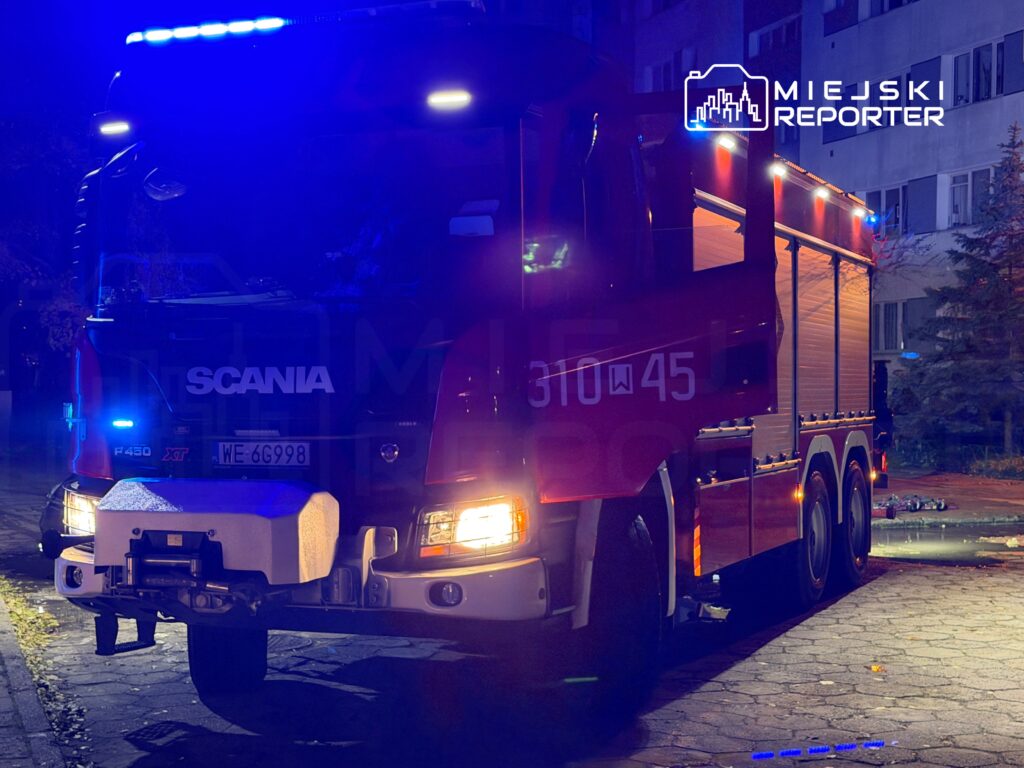 Czerwony wóz strażacki z włączonymi sygnałami świetlnymi stoi na oświetlonym chodniku w pobliżu budynku mieszkalnego.