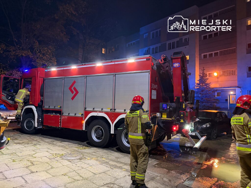 Strażacy w hełmach kontrolują sprzęt w pobliżu wozu strażackiego na oświetlonym placu, obok budynku mieszkalnego.