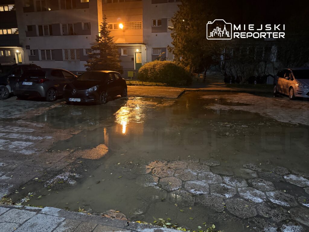 Zalany parking z kilkoma samochodami, w tle oświetlony budynek mieszkalny. Woda odbija światło lamp ulicznych.