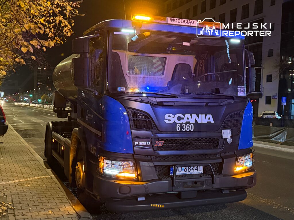 Ciężarówka marki Scania z oświetleniem sygnalizacyjnym zaparkowana na ulicy nocą, w tle widoczne iluminacje miejskie.