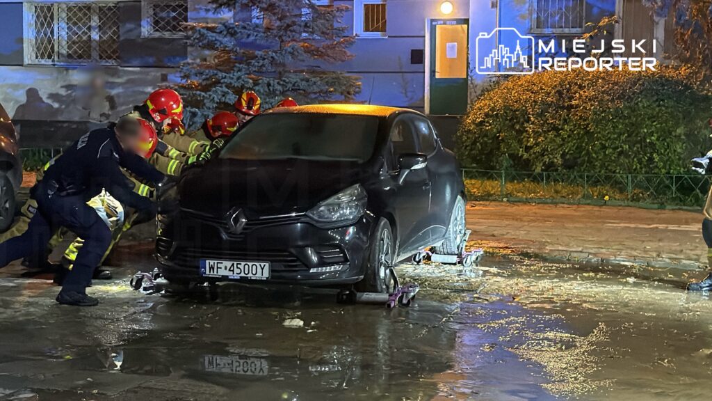 Grupa strażaków w kaskach wciąga samochód osobowy na wózkach transportowych na mokrej nawierzchni.