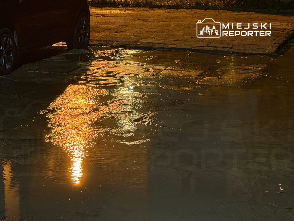 Czarne auto zaparkowane obok kałuży, w której odbijają się kolorowe światła uliczne.