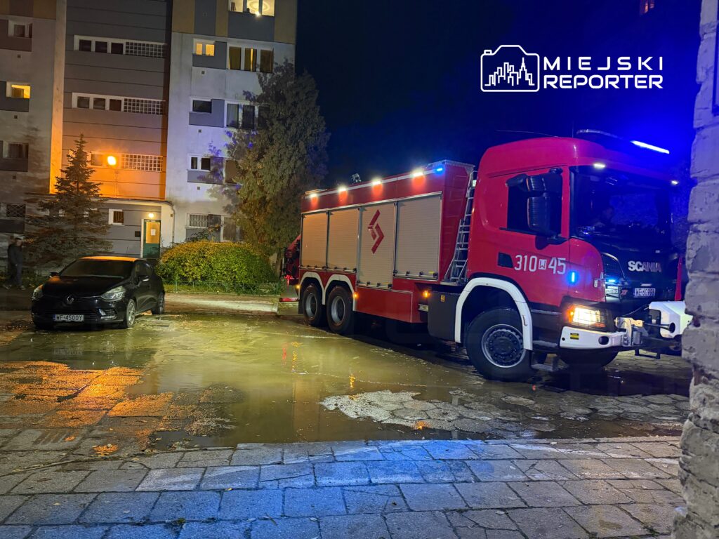 Czarny samochód osobowy zaparkowany obok wozu strażackiego na podjeździe budynku mieszkalnego w nocy.