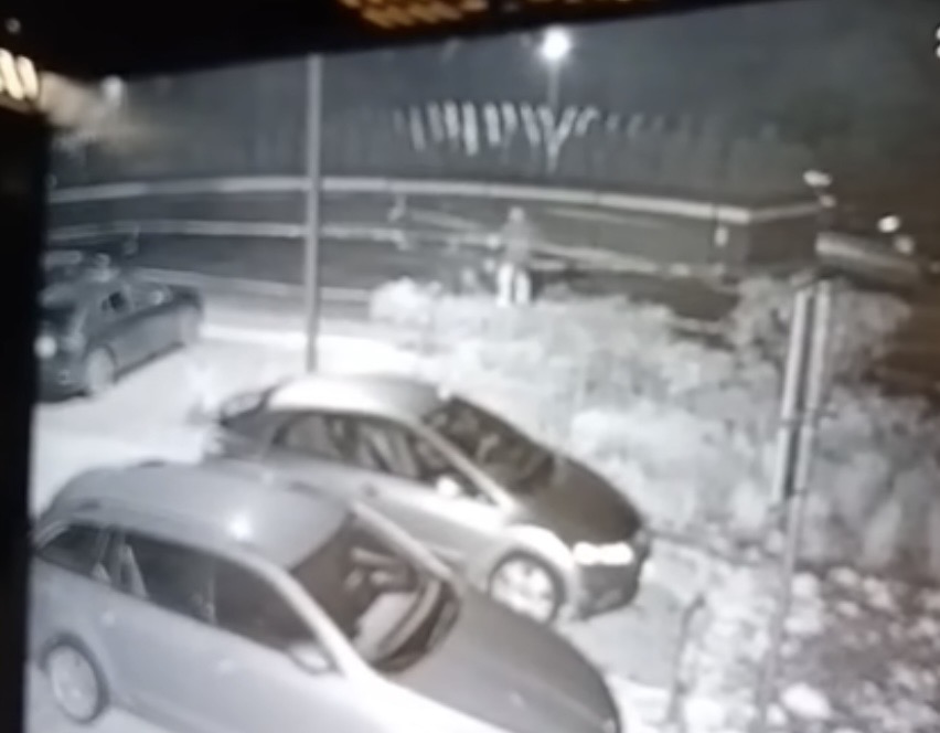 Mężczyzna w ciemnym ubraniu stoi obok zaparkowanych samochodów na oświetlonym parkingu nocą.