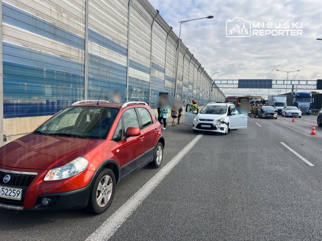 Czerwony samochód osobowy zatrzymany na drodze, obok uszkodzony pojazd, a w tle strażacy udzielający pomocy.