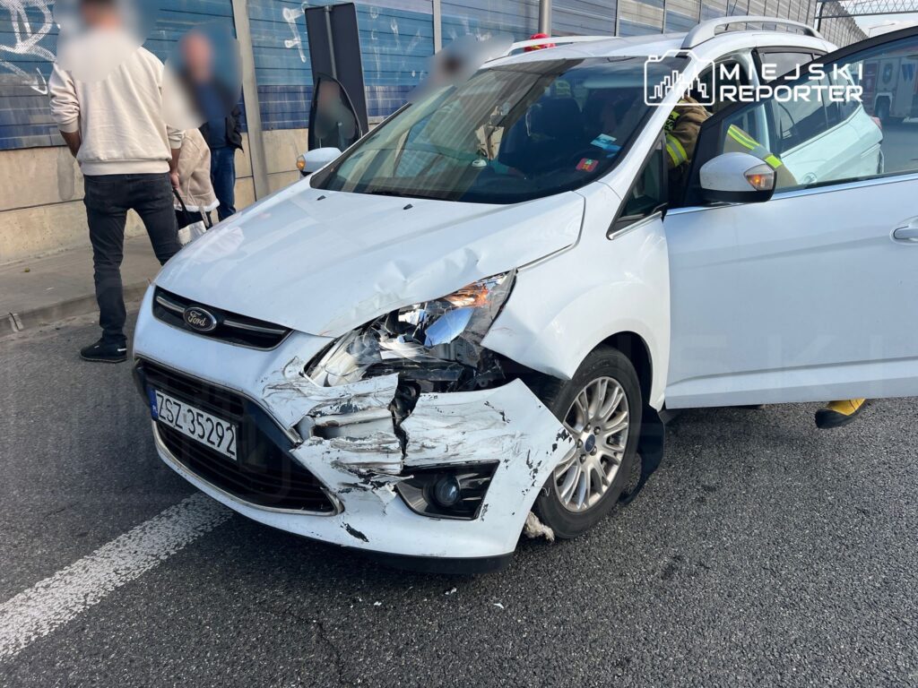 Biały samochód osobowy z uszkodzoną przednią częścią stoi na ulicy, obok grupka ludzi obserwuje sytuację.