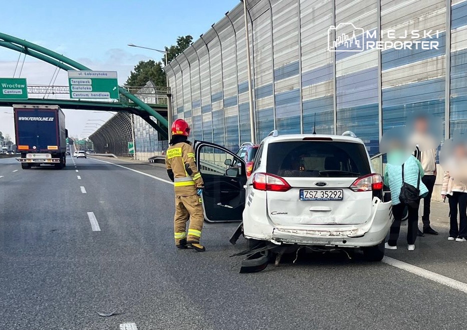 Strażak w mundurze rozmawia z osobami stojącymi obok uszkodzonego samochodu na drodze ekspresowej.