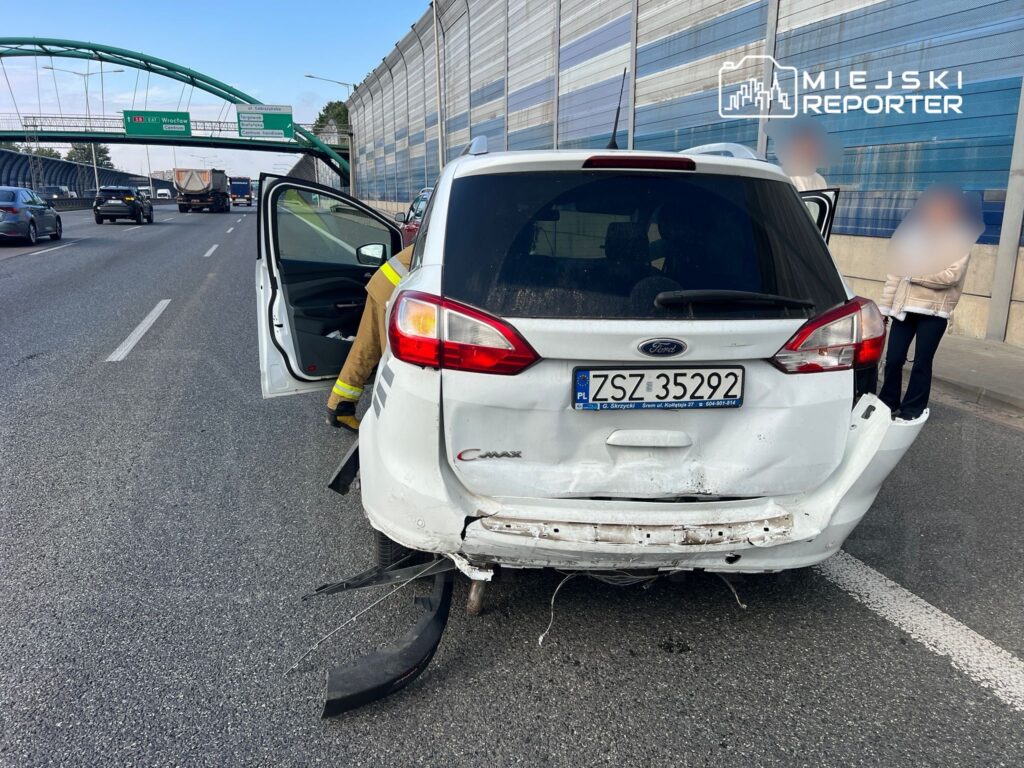 Biały samochód osobowy z uszkodzeniami tyłu stoi na pasie ruchu, obok dwóch osób rozmawiających.